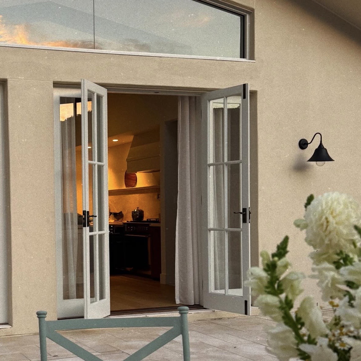 Exterior render at dusk, French doors open onto a warmly lit kitchen, wall sconce and white flowering shrub in the foreground