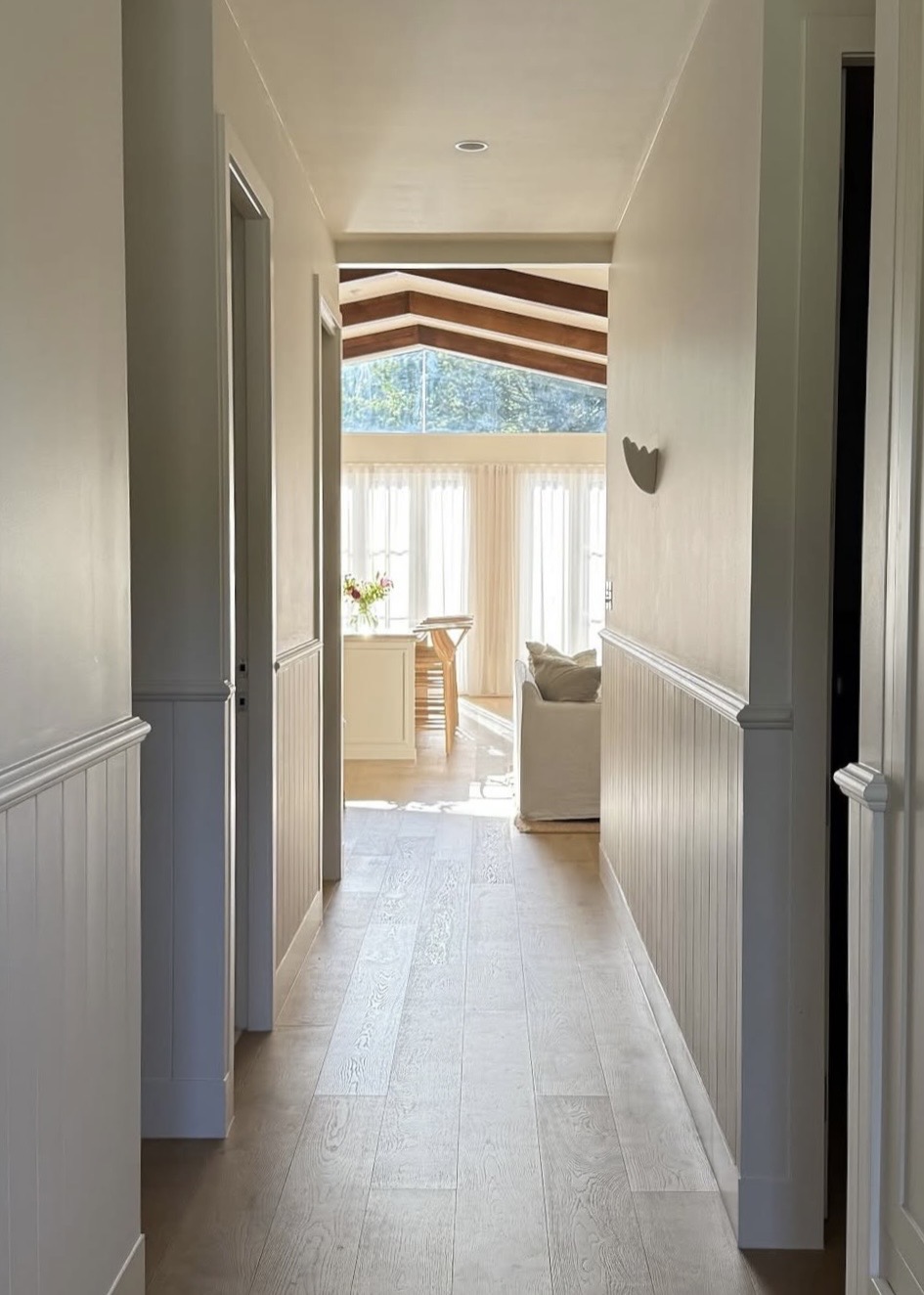 Beadboard-panelled hallway leading to a vaulted sunroom with timber beams, sheer curtains and a slipcovered armchair