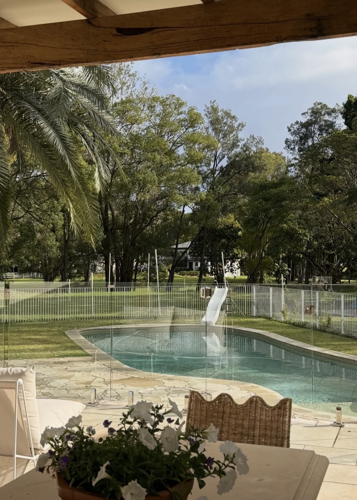 Pool with a slide and glass fencing, seen from under a timber pergola beam with a scalloped wicker chair and potted petunias