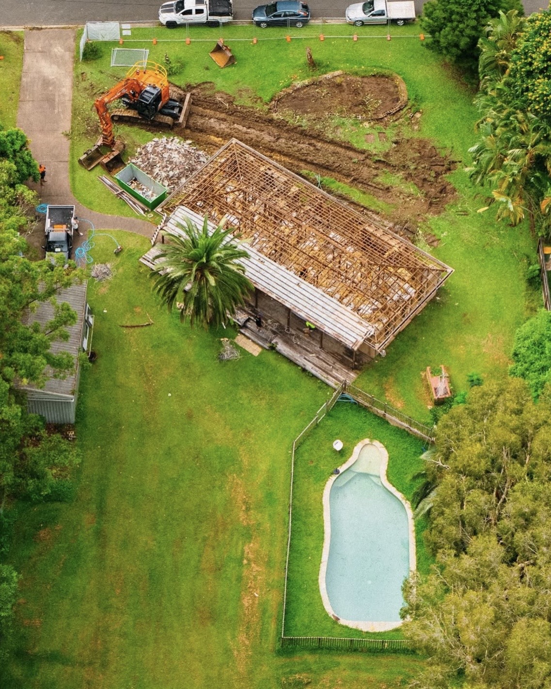 Aerial view of the property mid-construction — the original house stripped back to roof framing, an excavator, skip bin and the existing pool