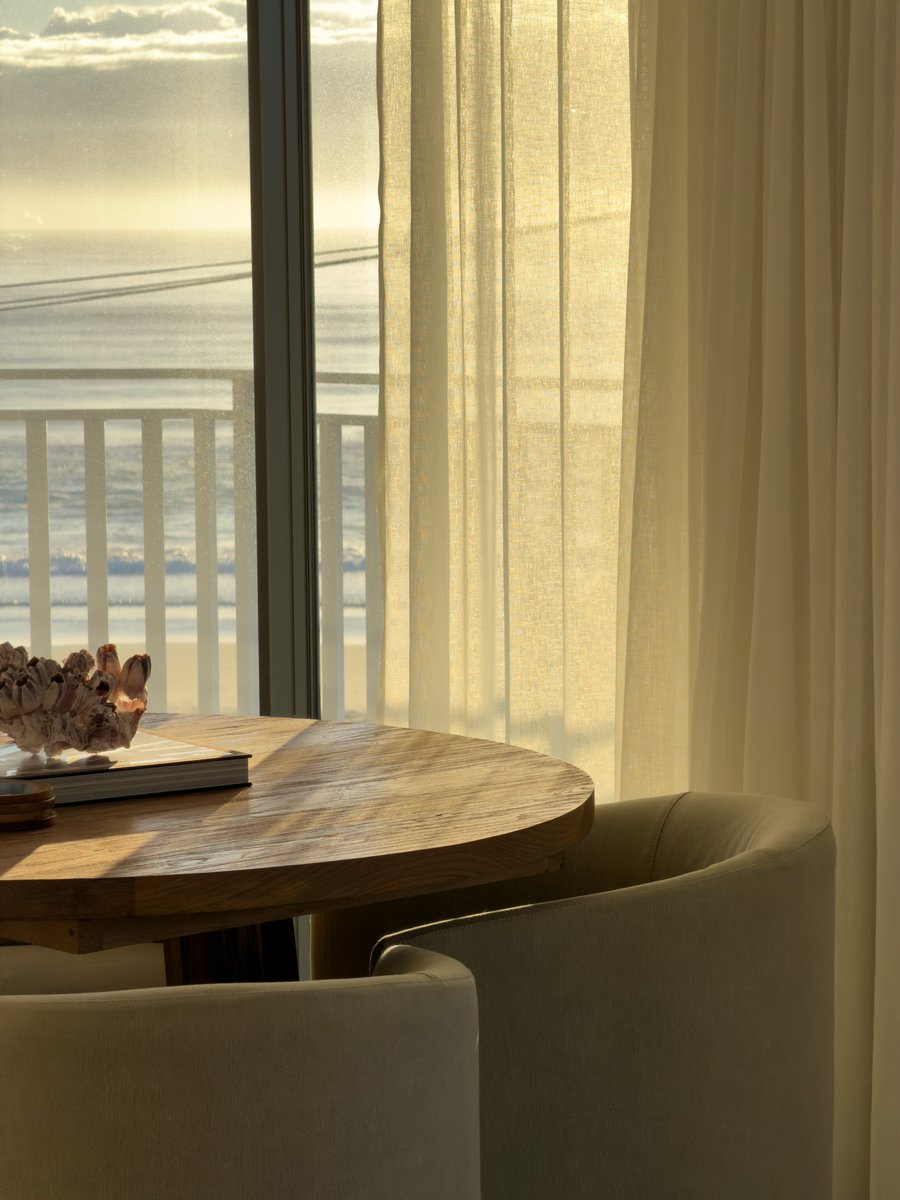 Round dining table styled with a coral ornament and books, cream leather chairs beside sheer curtains with a balcony and ocean view beyond