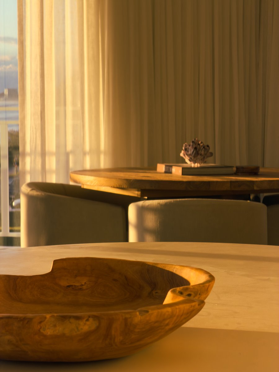 Carved timber bowl on a round dining table, with sheer curtains and a second table styled with books and a coral ornament beyond
