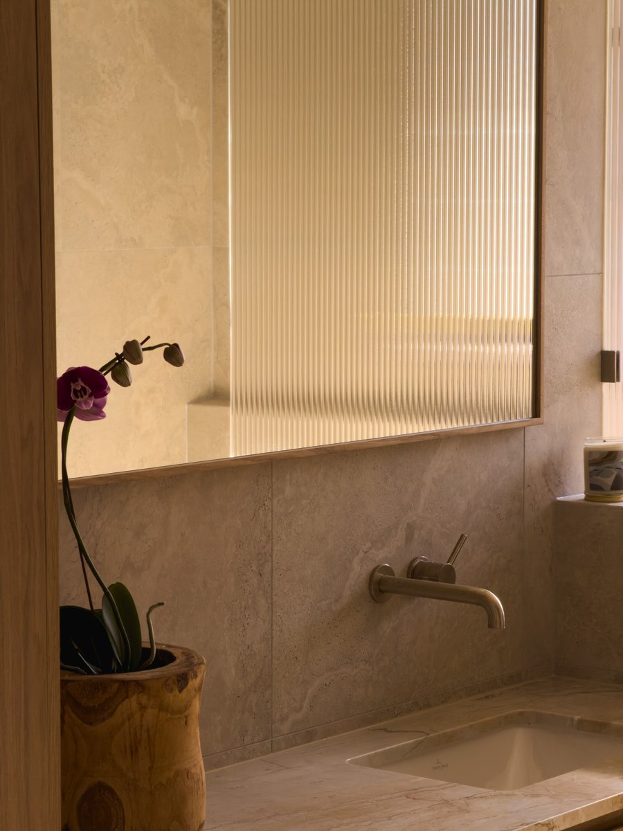 Bathroom vanity detail with a purple orchid, timber-framed mirror and wall-mounted tapware over a travertine benchtop