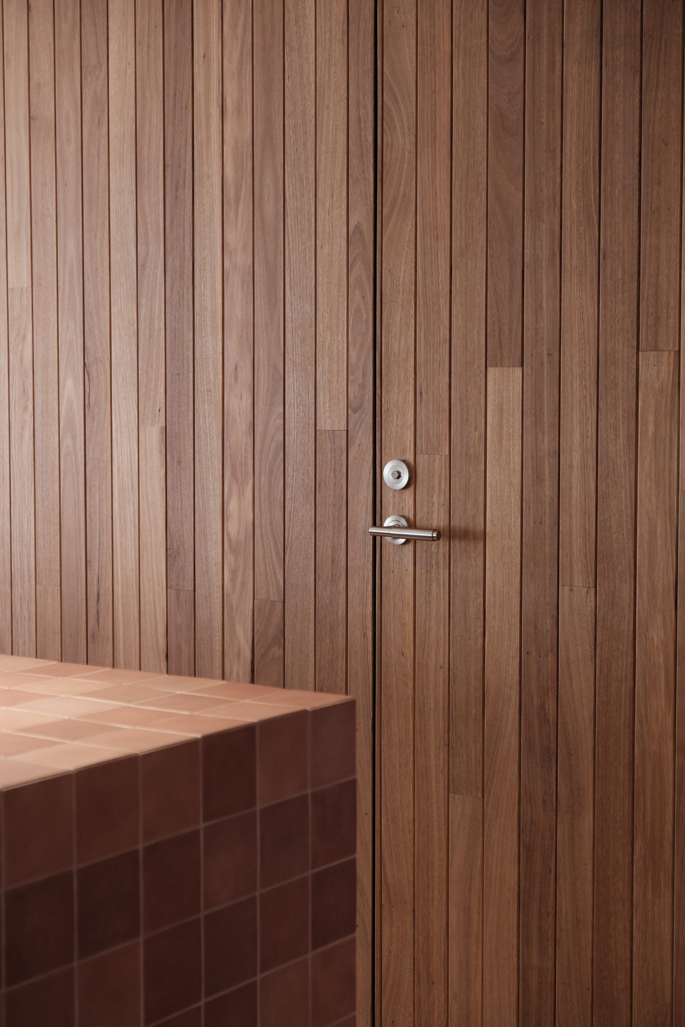 Timber-slat panelled door with a brushed-steel lever handle, next to a terracotta-tiled bench